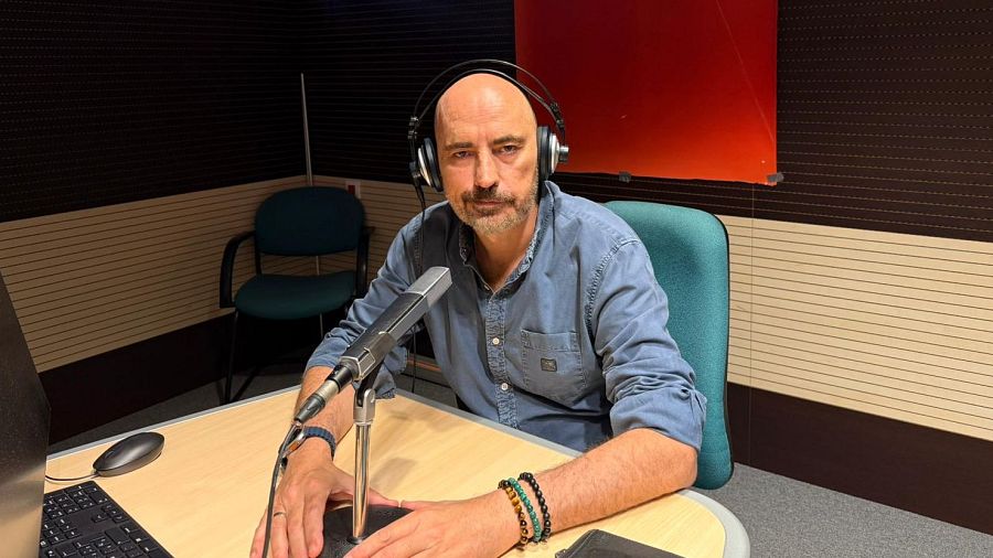 Periodista en cabina de radio con auriculares, micrófono, teclado y ratón.  Usa camisa azul y pulseras.  Paneles de madera clara y oscura en la pared.