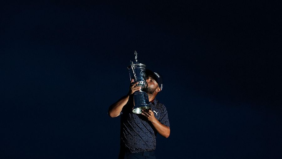 Golfista celebra victoria besando un trofeo; viste gorra y polo oscuro con lunares blancos.  Emoción y triunfo.
