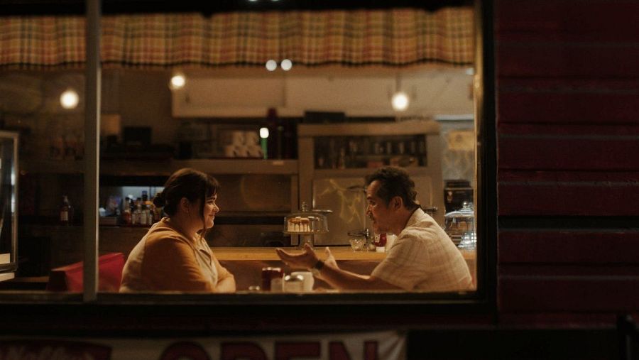 Escena de restaurante: hombre y mujer conversan animadamente a una mesa, con iluminación cálida y atmósfera íntima.  Se ve parte de la barra y un letrero 'OPEN'.