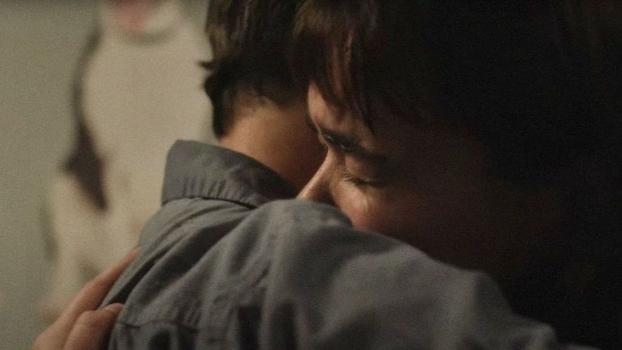 Primer plano de un abrazo entre dos personas; una con cabello oscuro abraza a otra con camisa gris.  Escena con fondo desenfocado.