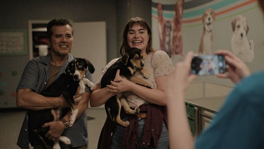 Pareja sonriente con cachorros, siendo fotografiados en un lugar con decoración de perros.  Escena de la película 