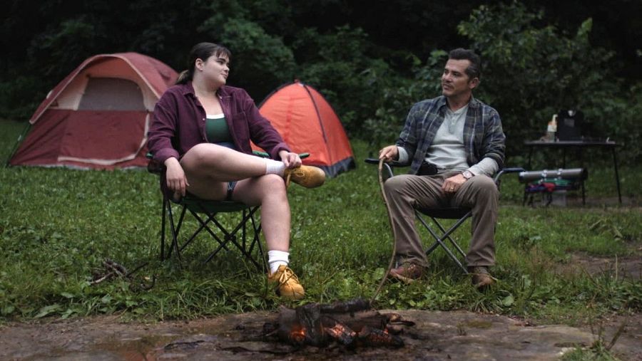 Dos personas, un hombre y una mujer, conversan junto a una fogata en un campamento con tiendas de campaña y mesa.