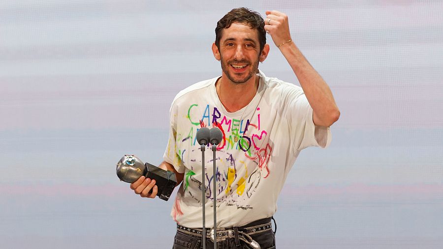 Actor recibiendo premio en escenario, sonriente y con el puño levantado.  Viste camiseta blanca con dibujos y cinturón con tachuelas.
