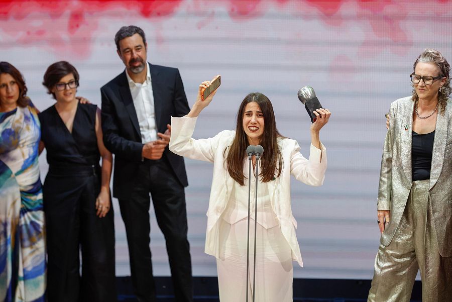 Directora teatral recibiendo premio en escenario, vestida de blanco, con expresión de alegría, sosteniendo el premio y un móvil.