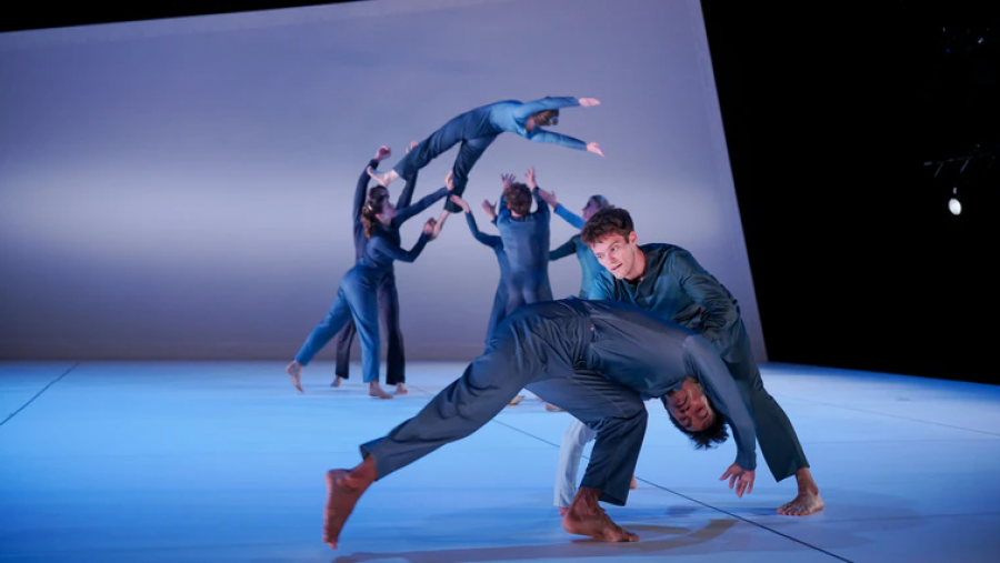 Coreografía dinámica de bailarines en monos azules, algunos sostenidos en el aire, con iluminación azul fría. Escena de 