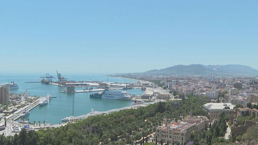 Panorámica de Málaga desde un punto elevado: puerto con barcos (incluido un ferry), ciudad y montañas al fondo bajo un cielo azul despejado.