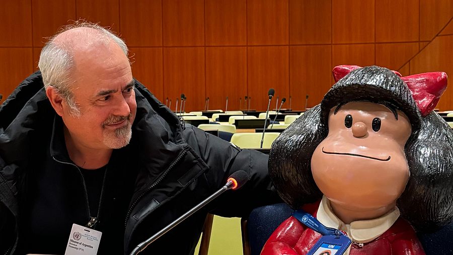 Quino junto a una figura de Mafalda en un auditorio. Lleva chaqueta negra y una credencial.  Mafalda viste su característico vestido rojo.