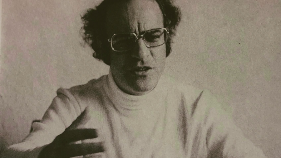 Imagen en blanco y negro de Quino joven, con gafas, cabello rizado y jersey de cuello alto; parece hablar o gesticular.