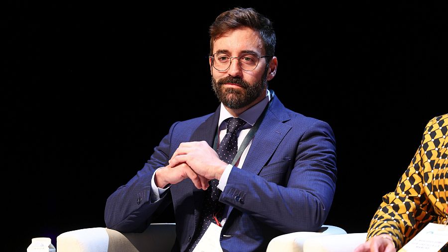 Jon Groizard, en traje azul marino y camisa blanca, sentado con expresión seria, mira hacia la izquierda.  Al fondo, parte de otra persona con ropa amarilla y negra.