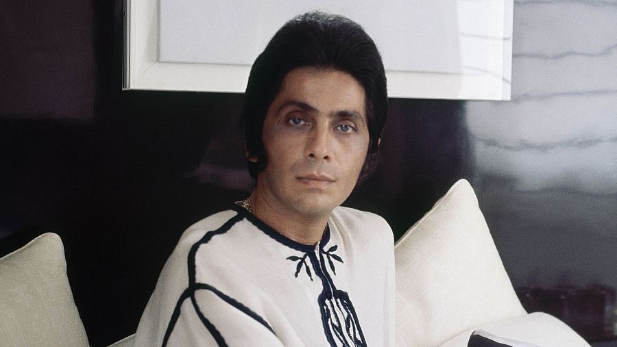 Fotografía de Valentino Garavani sentado en un sofá, con una camisa blanca bordada, en un ambiente elegante. 