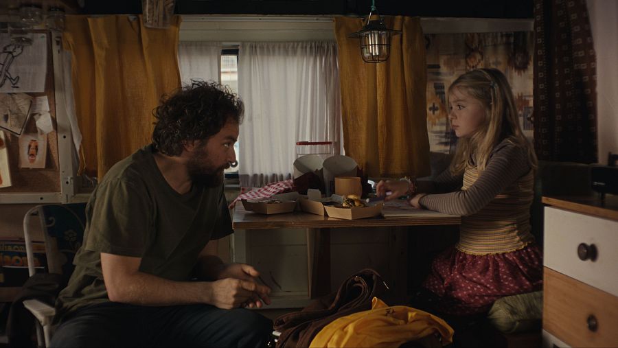 Hombre con barba y niña con vestido a rayas comparten comida en una caravana.  Envases de comida y dibujos en la pared. Ambiente acogedor.