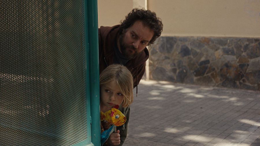 Hombre con barba y niña con juguete amarillo y azul se asoman con precaución desde detrás de una estructura metálica.  Su expresión denota miedo o alerta.