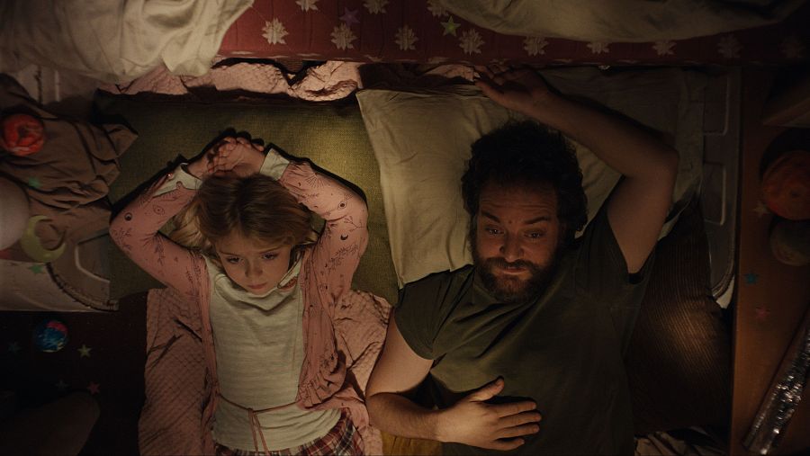 Padre e hija abrazados en cama dentro de una furgoneta decorada con motivos otoñales.  Ambiente nocturno y acogedor.