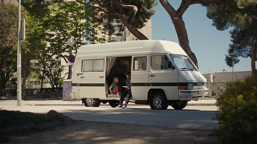 Escena de 'La furgo': una furgoneta blanca aparcada en una calle con árboles, con una niña y un hombre sentados en el escalón de la puerta lateral abierta.