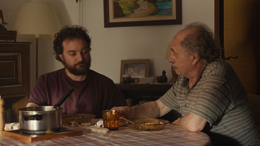 Escena hogareña: dos hombres, uno joven con barba y otro mayor con pelo canoso, comparten una comida en un comedor con mantel a cuadros.