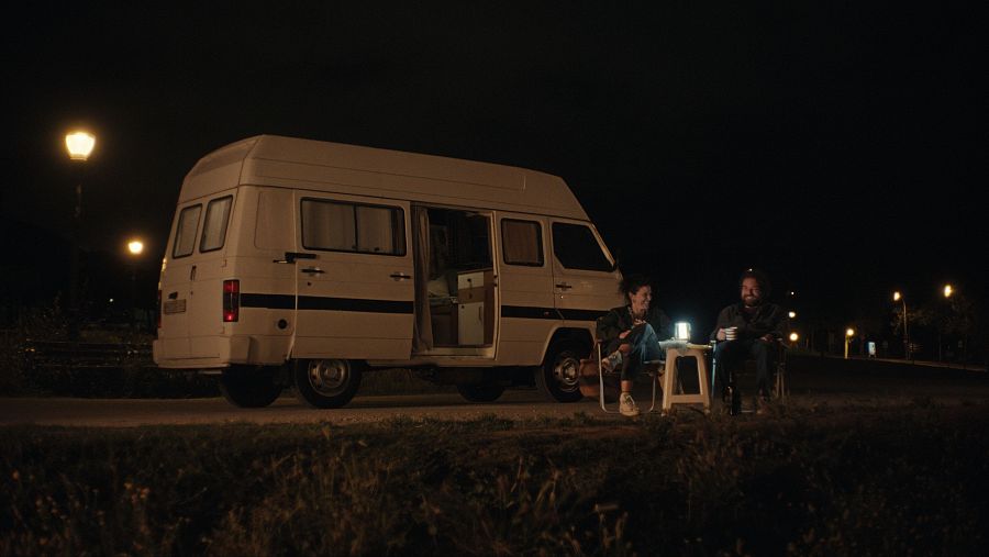 Parada nocturna junto a una carretera; una furgoneta blanca con puerta lateral abierta, dos personas relajadas sentadas cerca, disfrutando de la noche.