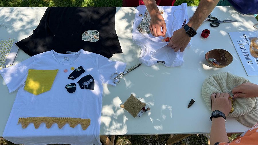 Taller de customización de camisetas con diversas herramientas de costura, telas estampadas y parches. Se ven camisetas blancas y negras decoradas.