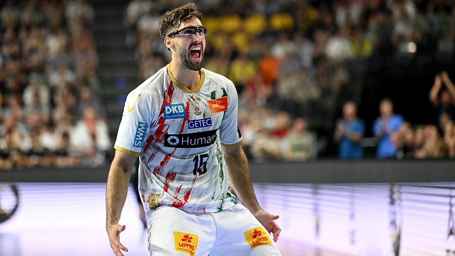 Jugador de balonmano celebrando un gol, con camiseta blanca con logos de patrocinadores y pantalones blancos con logo Lotto.  Expresión de euforia.