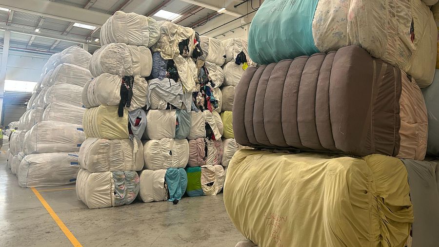 Fardos de textiles de diferentes tamaños y colores apilados en una nave industrial. Algunos fardos están envueltos en plástico transparente. Marcas amarillas en el suelo de cemento.