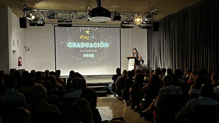 Graduación RTVE Instituto 2025.  Discurso de la directora ante los graduados en un auditorio iluminado, con pantalla mostrando el logo institucional y felicitaciones.