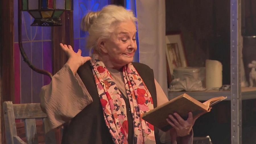 Escena teatral: Mujer mayor leyendo un libro, iluminada en primer plano; fondo con lámpara y estantería con objetos. Ambientación interior.