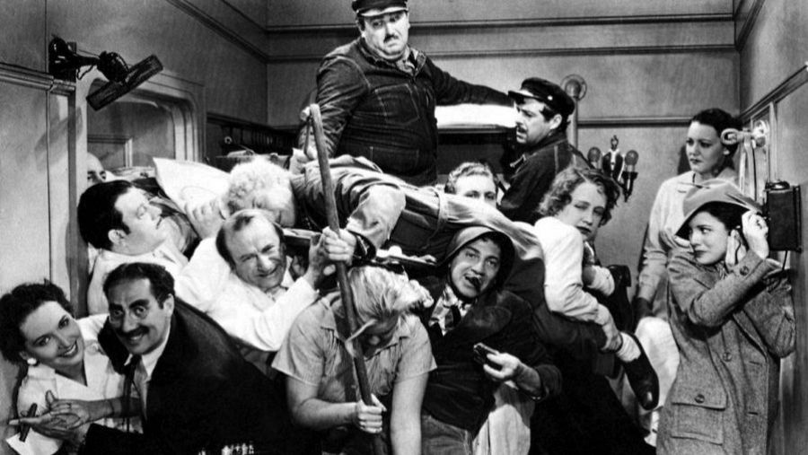 Caótico camarote repleto de gente; dos hombres uniformados intentan mantener el orden en una escena cómica en blanco y negro.