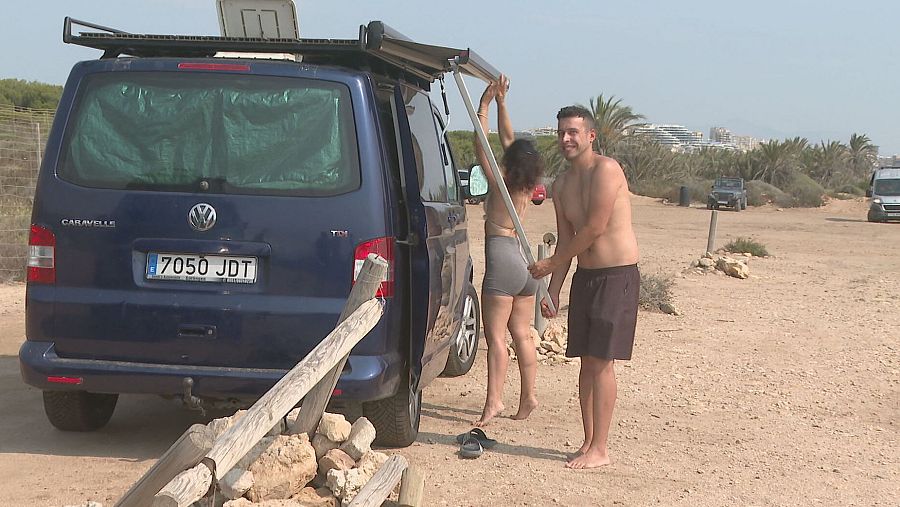 Pareja en bañador despliega toldo de furgoneta azul en playa arenosa.  Otros vehículos y vegetación al fondo.