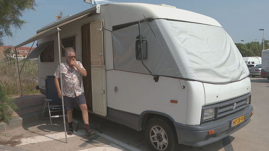 Hombre en ropa informal descansando junto a una autocaravana blanca con toldo.  Ambiente de camping o aparcamiento con otras caravanas.