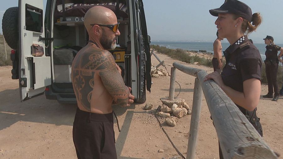 Agente de policía conversa con un hombre junto a una autocaravana blanca. El hombre, con tatuajes en los brazos, está cerca de la puerta abierta del vehículo.  Al fondo, se ve el mar y otros agentes.
