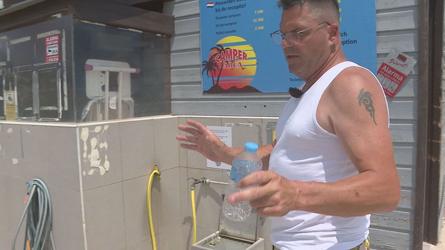 Hombre con camiseta blanca y gafas vaciando aguas negras en área de cámpers.  Mangueras y cartel informativo presentes.