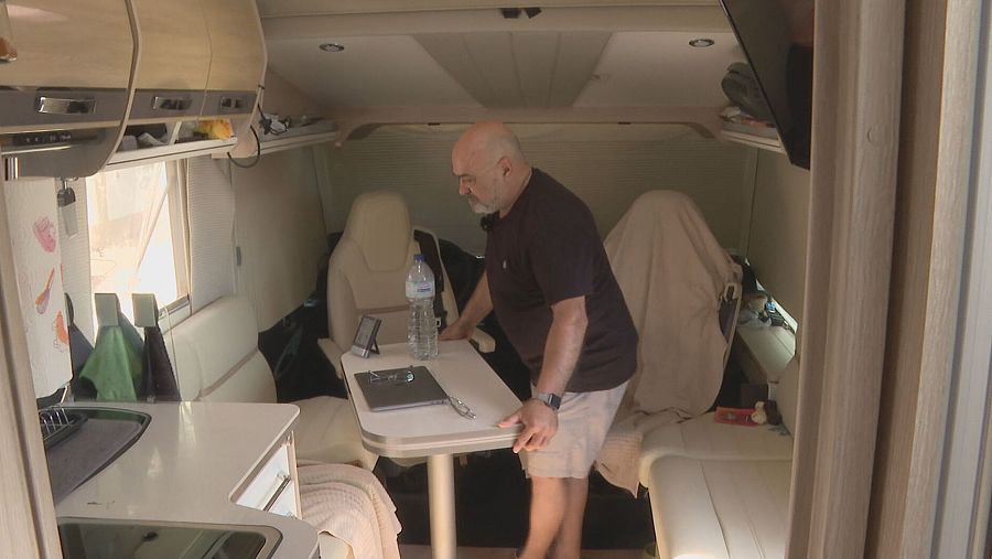 Interior de autocaravana con un hombre de pie junto a una mesa con una tablet y una botella de agua.  Armarios, televisión y asientos tapizados completan el espacio.