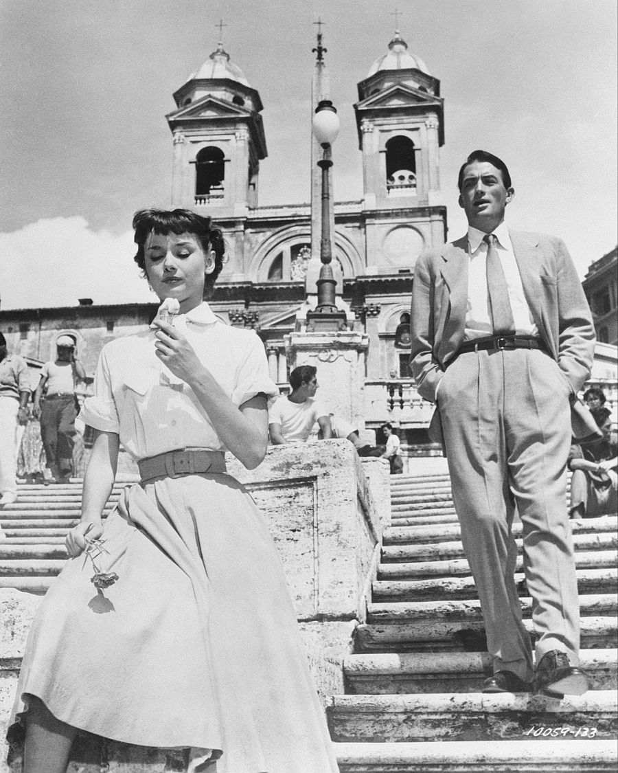 Audrey Hepburn, sentada en una escalinata, come un helado y sostiene una flor; Gregory Peck, de pie cerca, la observa.  Imagen en blanco y negro con atmósfera romántica.