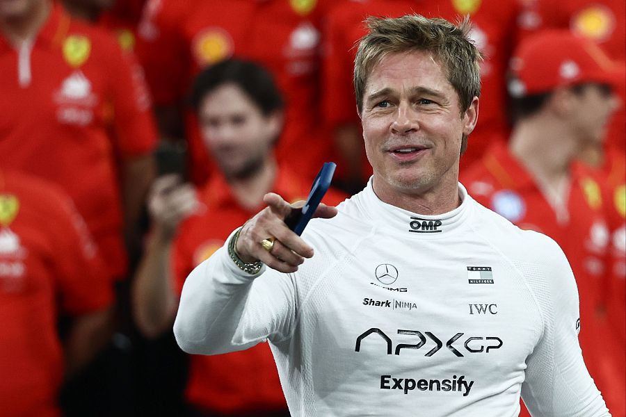 Actor grabando escenas en un circuito de carreras. Viste camisa blanca con logos y señala con un dedo mientras sostiene un objeto pequeño. Fondo con personal de equipo.