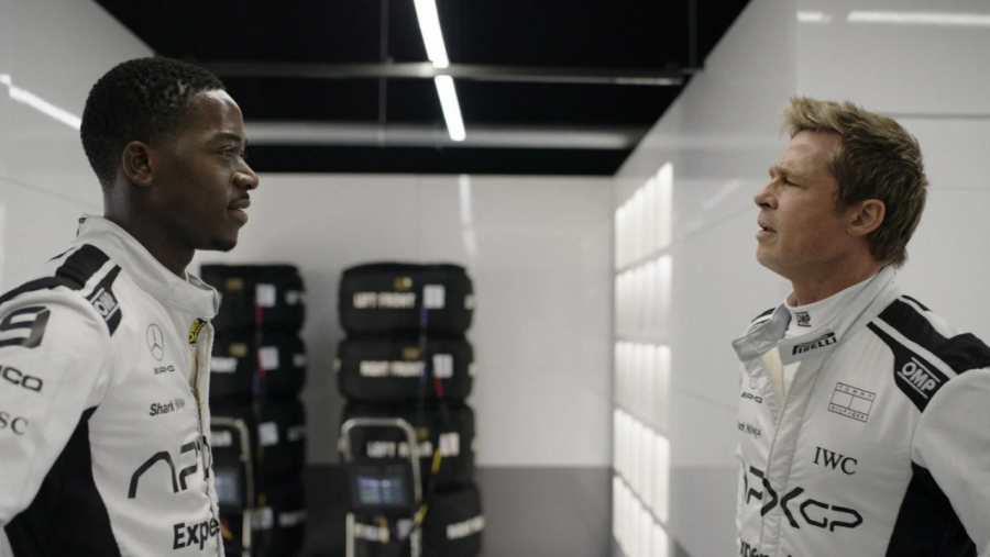 En el área de boxes, dos pilotos con monos blancos y detalles negros conversan.  Neumáticos apilados al fondo.