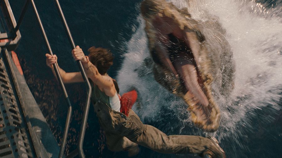 Mujer colgada de una escalera sobre agua, amenazada por un gran reptil que emerge.  Acción y peligro inminentes.