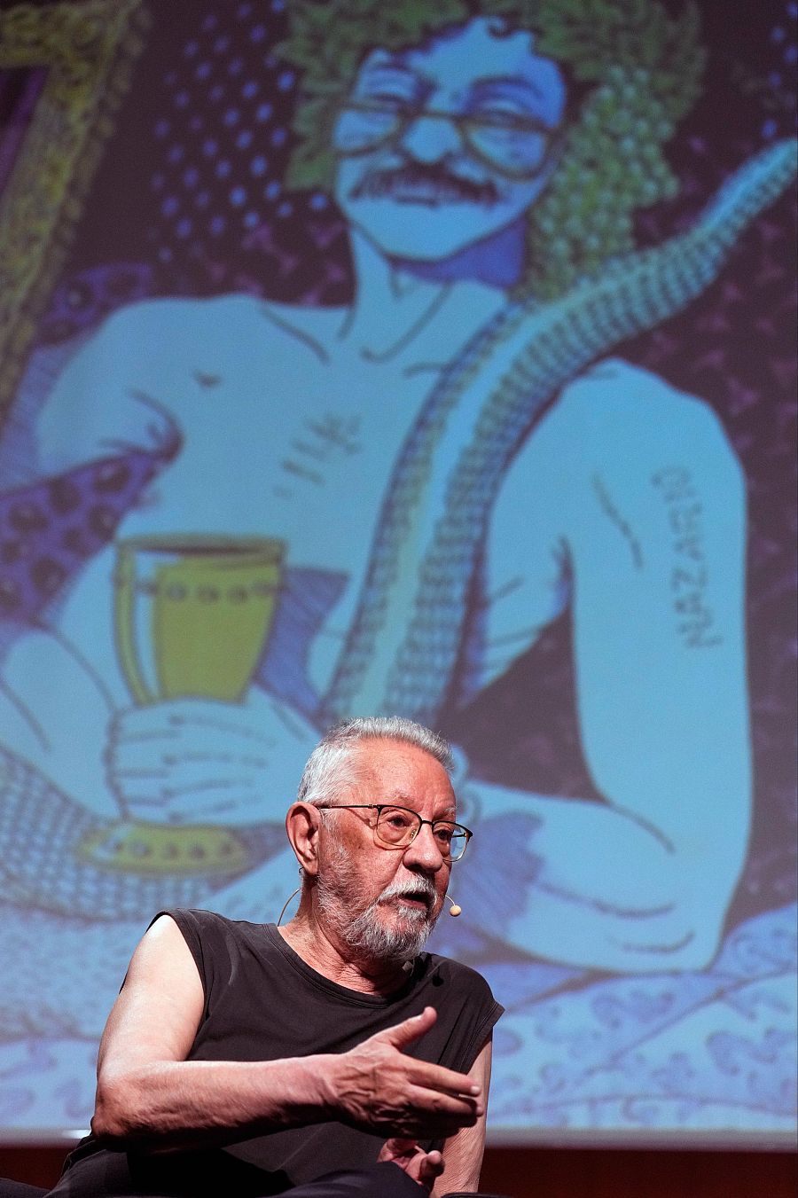 Historietista con gafas y barba canosa sentado, gesticulando.  Detrás, proyección de un personaje de cómic con gafas y bigote, con firma visible.