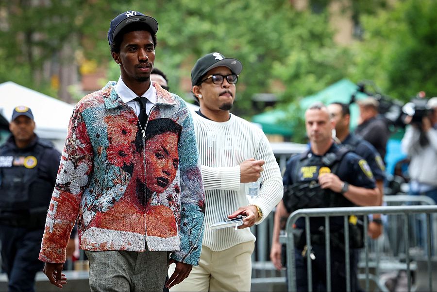 Padre e hijo llegan a un juicio en Nueva York. Él viste jersey blanco y gafas; él, chaqueta estampada, pantalones grises y gorra.