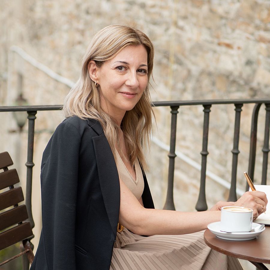 Escritora sentada en terraza con taza de café y libreta, escribiendo con bolígrafo dorado. Viste chaqueta negra y blusa beige.