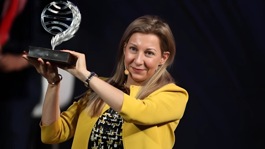Mujer de pelo rubio y chaqueta amarilla, sosteniendo con alegría un premio circular con hojas sobre base rectangular.