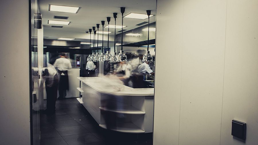 Cocina profesional con isla central blanca, lámparas colgantes y cocineros en uniforme trabajando activamente. Ambiente luminoso y dinámico.