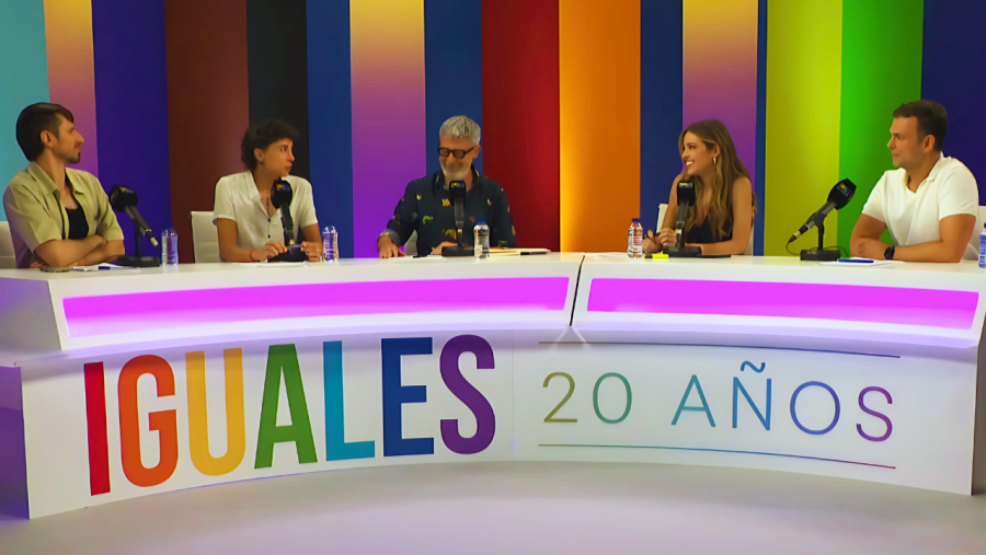 Mesa semicircular blanca con franja rosa, cinco personas con micrófonos y agua, fondo multicolor y letrero 'IGUALES 20 AÑOS'.