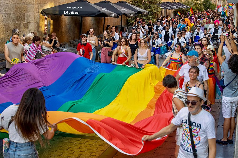 Manifestación del Orgullo LGTBIQ+: multitud portando una gran bandera arcoíris en una calle, ambiente festivo y reivindicativo.
