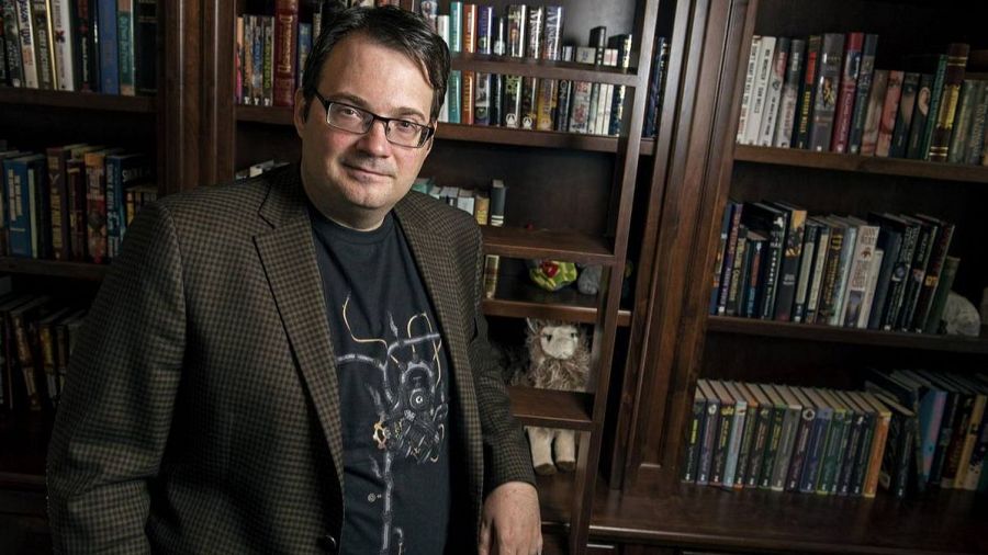 Brandon Sanderson, escritor, posa con gafas y camisa negra ante una estantería llena de libros y un pequeño peluche.