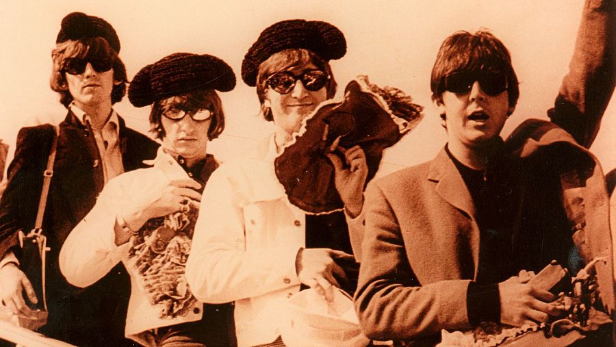 Los Beatles, con boinas y gafas de sol, llegan a España en 1965.  Imagen sepia mostrando a los cuatro miembros de la banda sosteniendo objetos.