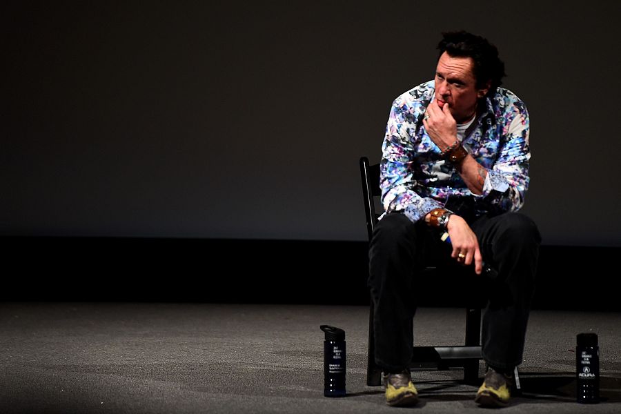 Actor sentado en silla, camisa floral, pantalones oscuros y botas verdes. Expresión pensativa, dos botellas de agua a sus pies. Fondo oscuro.