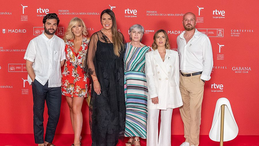 Presentación de premios de moda: siete personas posan en alfombra roja con photocall mostrando logotipos de patrocinadores.  Ambiente formal y festivo.