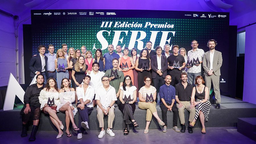 Premiados de la III edición de los Premios Seriemanía posando con sus trofeos en forma de 'M' en un escenario con iluminación violeta y pantalla de fondo.