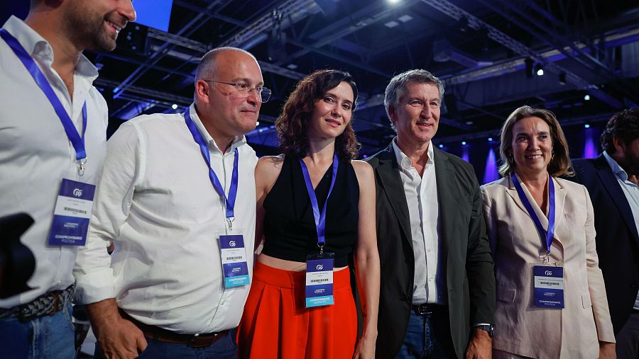 Isabel Díaz Ayuso, Alberto Núñez Feijóo y Cuca Gamarra, junto a otras personas con credenciales, en un evento público. Todos miran hacia adelante.