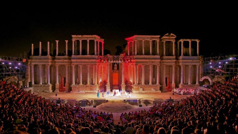 Espectacular vista nocturna del Teatro Romano de Mérida durante una representación. Artistas en escena, público en las gradas y una atmósfera vibrante.