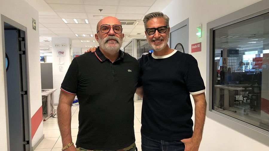 Dos hombres, uno con polo negro y pantalón de camuflaje, el otro con camiseta azul, conversan en un pasillo con el logo de RNE visible.  Se observa el cartel 